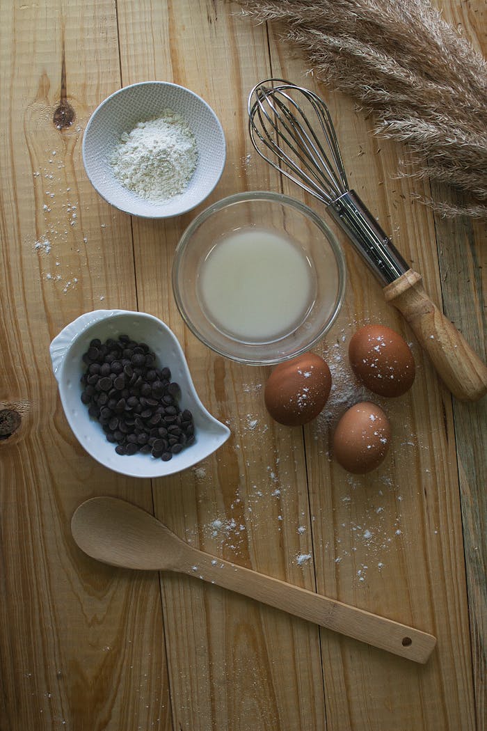 A flat lay of baking essentials including eggs, flour, chocolate chips, and a whisk on a wooden surface.